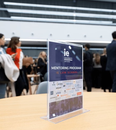A mentoring program sign displayed on a table at a crowded event in a modern lounge area.