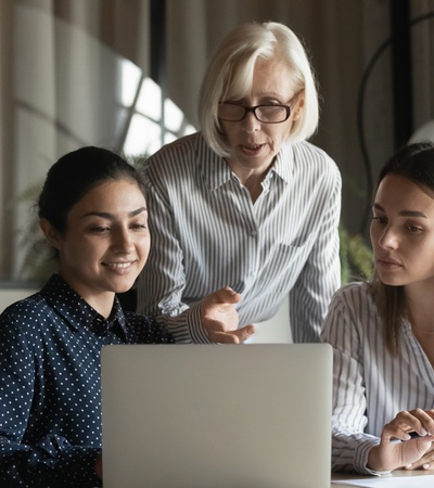 An older woman instructs two younger women using a laptop in an office setting.