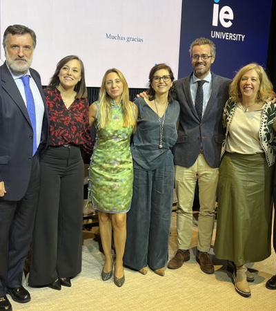 A group of seven adults are smiling and posing together at a public event in front of a stage with 'IE University' branding.