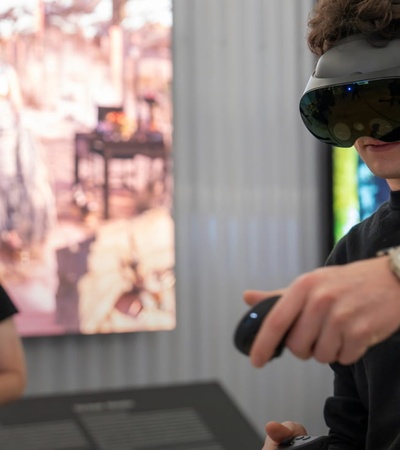 A man in the foreground uses virtual reality equipment while another man in the background watches in a room with artistic imagery.