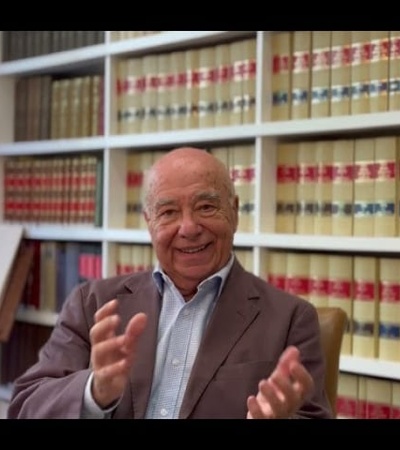 An elderly man smiling and gesturing with his hands in front of a bookshelf filled with binders and books.