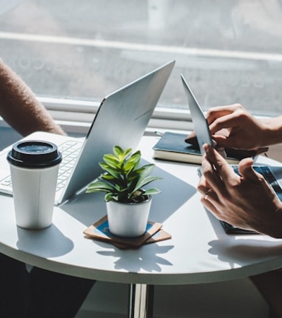 Two people working on laptops and tablets at a small table with a coffee cup and a plant.