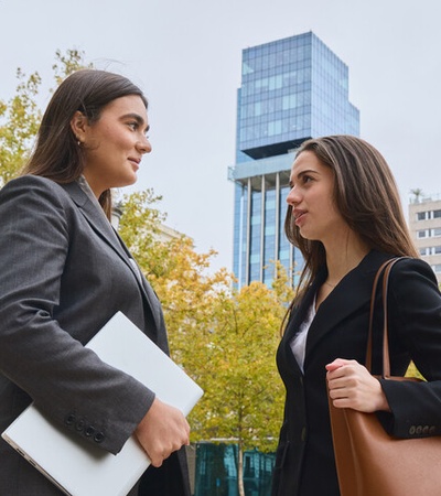 Two women are having a conversation while standing in an urban area with buildings in the background.