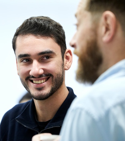 A young man with a beard smiling at another person during a conversation.