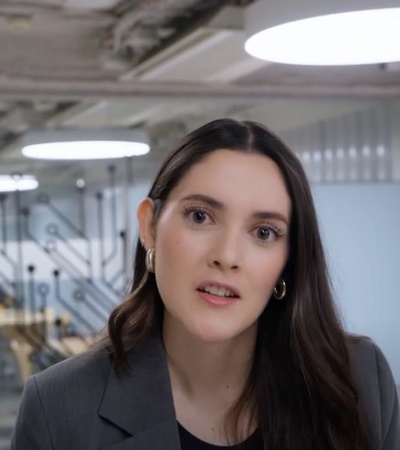 A woman in a professional attire stands in a modern office environment with soft overhead lighting.