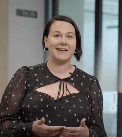 A woman in a patterned dress is speaking and gesturing with her hands in an office environment.