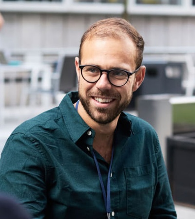A man in glasses smiling during a conversation with another person at an outdoor table.