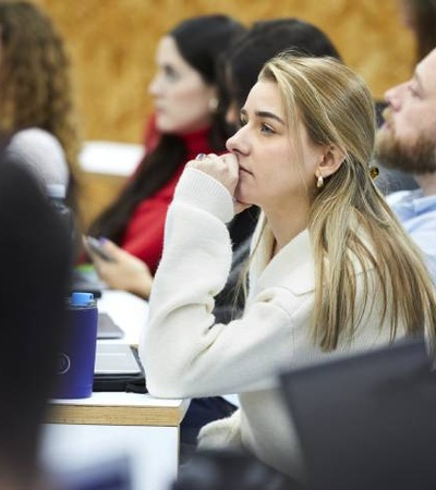 A woman in a white coat appears thoughtful while attending a seminar with other participants.