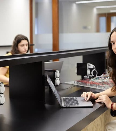 A young woman working on her laptop at a laboratory desk while another woman works in the background.