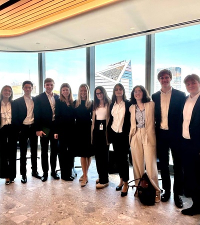 A group of ten business professionals in formal attire standing in a modern office environment with large windows and city view.