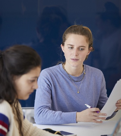A woman is attentively looking at another woman seated at a table with papers and a laptop.