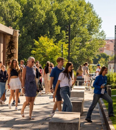 Students walking and socializing on a university campus on a sunny day, with a store and trees in the background.