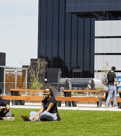 Two people are sitting on a grassy area in an urban park next to a modern building.