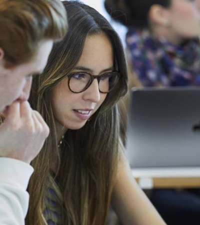 Two students, a male and female, are focused on a laptop screen while sitting at a table in a classroom.
