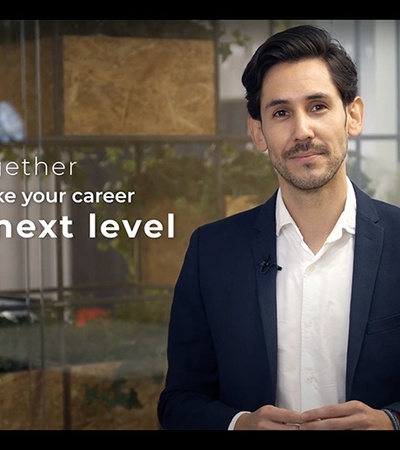 A man in a suit stands indoors with a message 'Together we can take your career to the next level' displayed behind him.