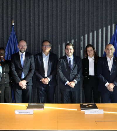 A group of eight people, four women and four men, stand behind a conference table with national flags in the background.