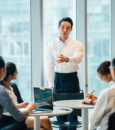 A man stands presenting to colleagues at a round table in a brightly lit office.