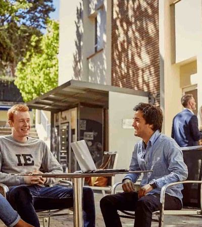Three students are having a discussion at an outdoor seating area of a campus, with other individuals passing by.