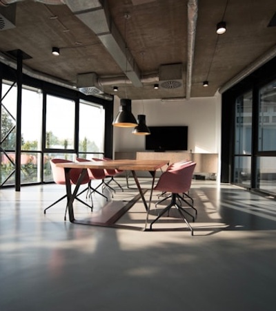 A modern conference room with large windows and a wooden table surrounded by pink chairs.