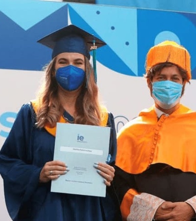 A female graduate in a blue gown and mask holds her diploma standing next to a person in an orange academic robe and mask during a graduation ceremony.