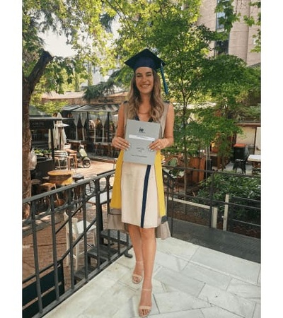 A young woman in a graduation cap and gown holding a diploma stands on a balcony with a restaurant background.