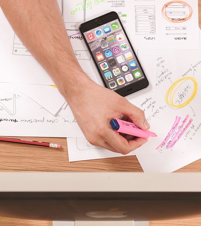 An overhead view of a person's hands working on a creative project with papers, pens, a smartphone, and stationery items on a wooden desk.