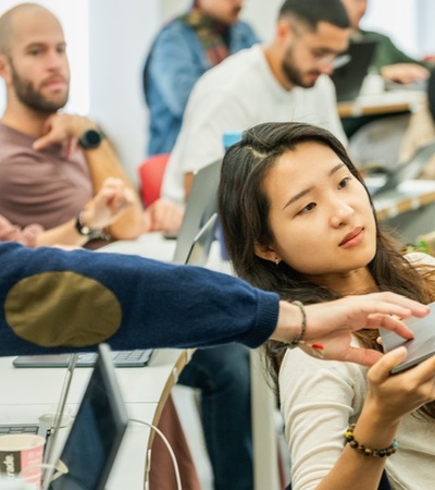 A student in a classroom is showing their laptop screen to another student.