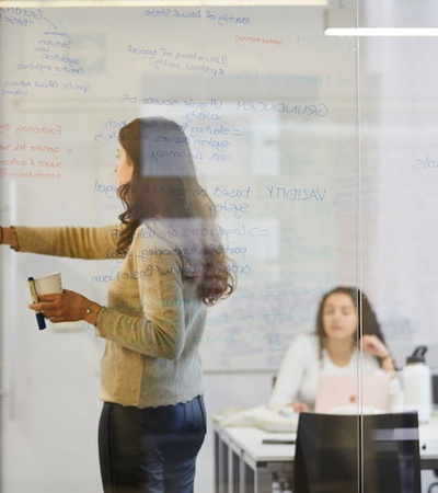 A woman is presenting ideas on a glass board while others listen in a modern office environment.
