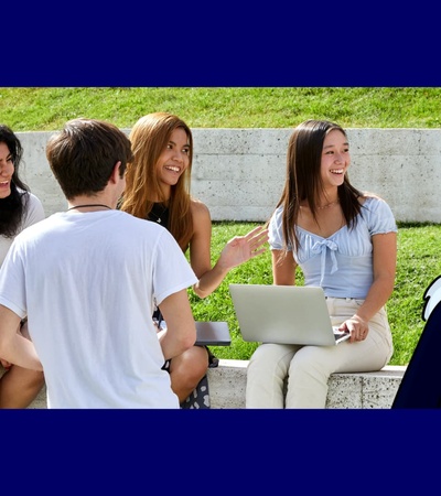 A group of five diverse students sitting outdoors on campus steps, engaging in a discussion with one student using a laptop.