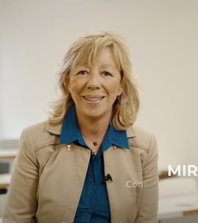 A woman smiling in a classroom environment, standing at the front with a caption identifying her as 'MIRIAM BIEGER'.