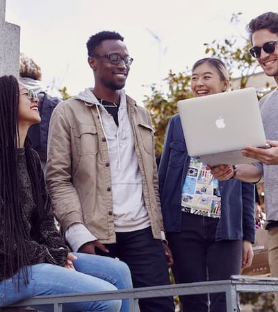 A group of young adults are smiling and interacting outdoors, one of them holding a laptop.