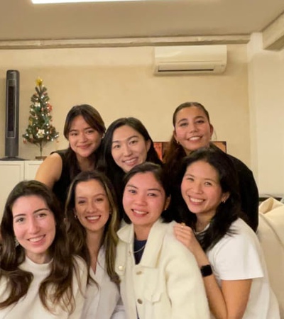 A group of seven women smiling in a cozy room with soft lighting and a small Christmas tree in the background.