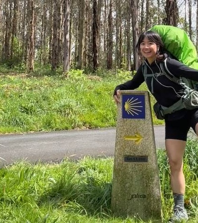 A smiling person in hiking gear poses playfully next to a signpost on a trail.