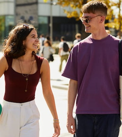 A young man and woman are walking and smiling together in a sunny, outdoor setting, with the woman holding a volleyball.