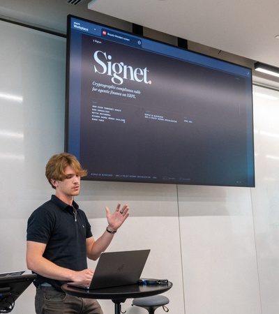 Three individuals are presenting in front of a large screen displaying the Signet platform.