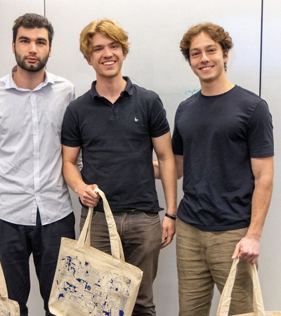 Three young men are standing together holding tote bags, smiling at the camera.