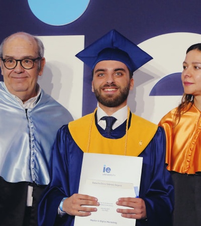 A graduate in a blue and yellow cap and gown holding a diploma, flanked by two adults in academic regalia.