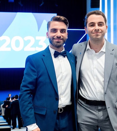 Two men in business suits smiling at a conference with a 'B+ 2023' banner in the background.