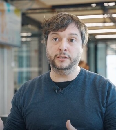 A man in a dark shirt is talking and gesturing with his hands in an office environment.