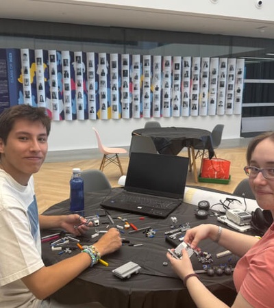 Two people sitting at a table with electronic components and a laptop, working on a project in a room with posters on the wall.