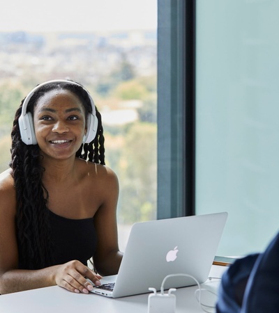 A young woman with headphones smiles while working on a laptop in a modern office space.
