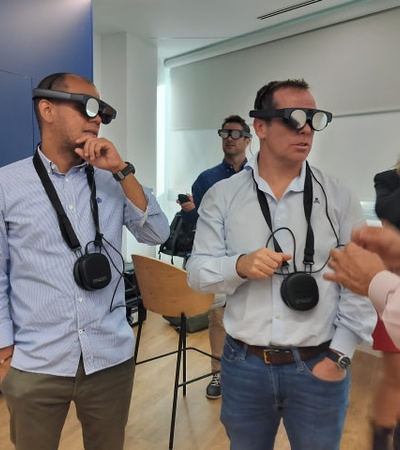 Two men wearing virtual reality headsets and holding cameras are interacting with another man in an indoor setting.