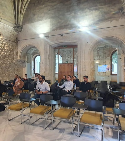 A speaker presents to an audience in a spacious, historic room with stone walls.