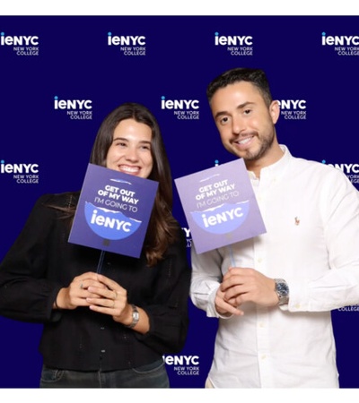Two smiling individuals hold signs in front of a blue background with text promoting ieNYC.