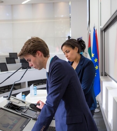 Two individuals are standing at a podium in a conference room, with flags in the background.
