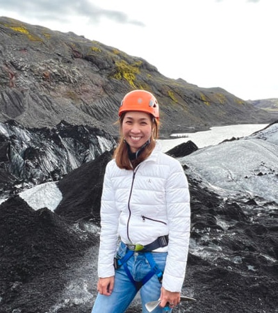 A woman in a white jacket and orange helmet stands smiling in a rugged mountainous landscape.