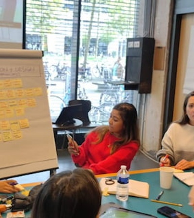 A group of diverse people engaged in a discussion around a table with notes and a whiteboard in a bright room.