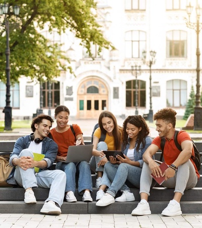 A group of five young adults happily using a smartphone while sitting together on steps in a campus setting.