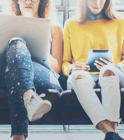 Five young adults sitting on a bench, each engaged with different digital devices.