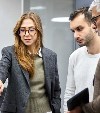 A young woman in glasses and a blazer is discussing something with two male colleagues in an office environment.